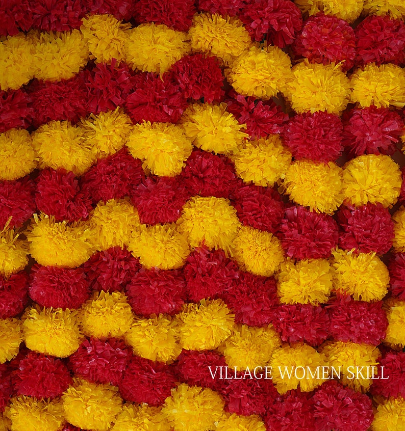May include: A close-up of a decorative garland made of red and yellow fabric flowers. The garland is arranged in a wavy pattern. The text "VILLAGE WOMEN SKILL" is visible at the bottom of the image.