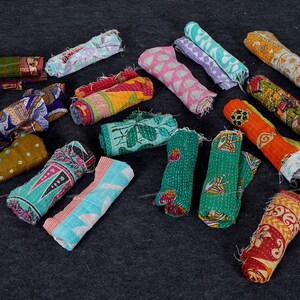 May include: A collection of colorful kantha quilts rolled up and arranged on a dark gray surface. The quilts are made of various fabrics and feature different patterns and designs.