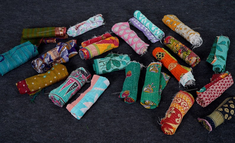 May include: A collection of colorful kantha quilts rolled up and arranged on a dark gray surface. The quilts are made of various fabrics and feature different patterns and designs.