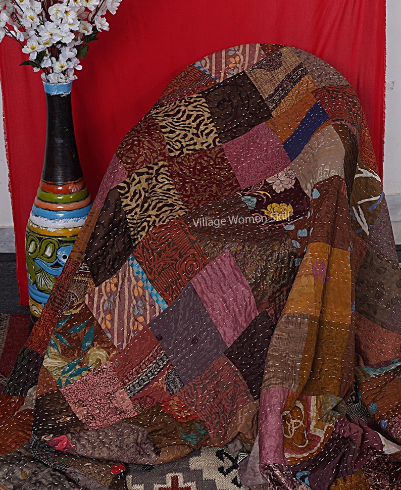 May include: A patchwork quilt made from various fabrics in shades of brown, pink, and blue. The quilt is folded and draped over a chair. The text "Village Women Skill" is visible on the quilt.