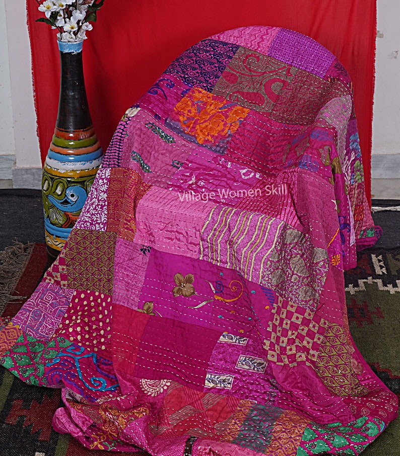 May include: A pink and purple patchwork quilt made from recycled saris. The quilt is folded and draped over a chair. The quilt has a variety of patterns and textures, including floral, geometric, and paisley.