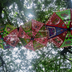 May include: Three colourful, patterned umbrellas hang from tree branches. The umbrellas are decorated with embroidered elephants, flowers, and mirrors. The umbrellas are pink, green, blue, and red.