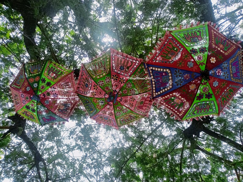 May include: Three colourful, patterned umbrellas hang from tree branches. The umbrellas are decorated with embroidered elephants, flowers, and mirrors. The umbrellas are pink, green, blue, and red.