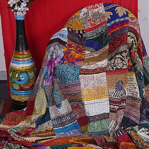 May include: A colorful patchwork quilt made from recycled saris. The quilt is made up of squares of different fabrics, each with a unique pattern and color. The quilt is draped over a chair and has a rustic, vintage look.