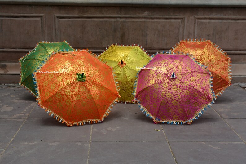 K&ouml;nnte beinhalten: F&uuml;nf dekorative, offene Regenschirme. Sie zeigen ein goldenes Blumenmuster auf orangefarbenem, gr&uuml;nem, gelbem und pinkfarbenem Hintergrund. Die R&auml;nder sind mit dekorativen Fransen versehen, was sie besonders macht.