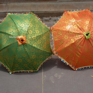 K&ouml;nnte beinhalten: Zwei dekorative Sonnenschirme, einer gr&uuml;n und einer orange, mit goldenen Blumenmustern. Jeder Sonnenschirm hat eine dekorative Fransen und einen zentralen floralen Akzent. Die Sonnenschirme sind auf einer Steinoberfl&auml;che ge&ouml;ffnet.