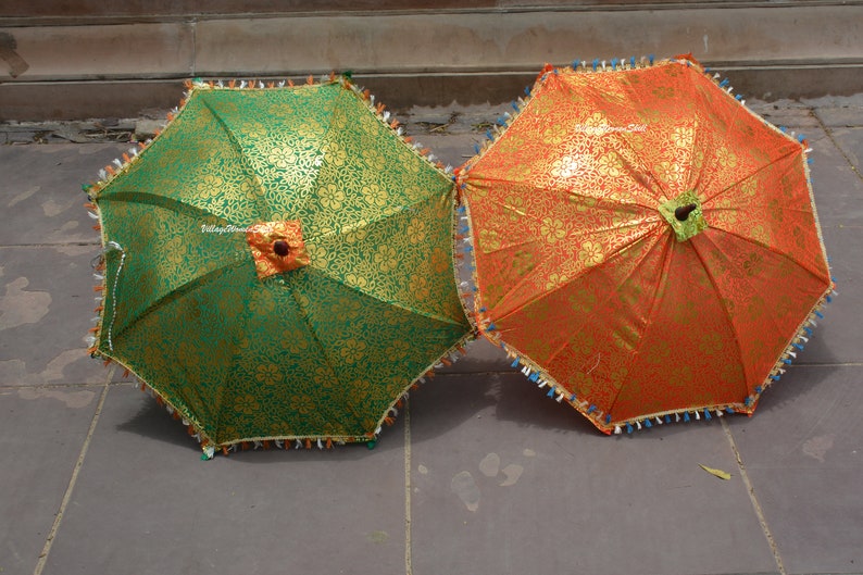 K&ouml;nnte beinhalten: Zwei dekorative Sonnenschirme, einer gr&uuml;n und einer orange, mit goldenen Blumenmustern. Jeder Sonnenschirm hat eine dekorative Fransen und einen zentralen floralen Akzent. Die Sonnenschirme sind auf einer Steinoberfl&auml;che ge&ouml;ffnet.