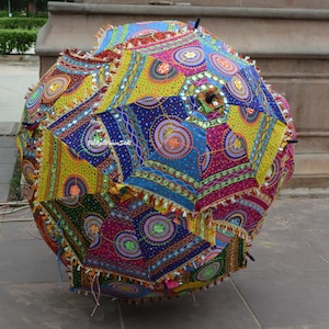 May include: A large, colorful, patchwork umbrella with a vibrant design. The umbrella features a variety of geometric shapes in shades of blue, yellow, green, and pink, adorned with decorative tassels. The umbrella is open and resting on a stone surface.