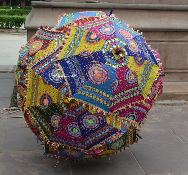 May include: A large, colorful, patchwork umbrella with a vibrant design. The umbrella features a variety of geometric shapes in shades of blue, yellow, green, and pink, adorned with decorative tassels. The umbrella is open and resting on a stone surface.