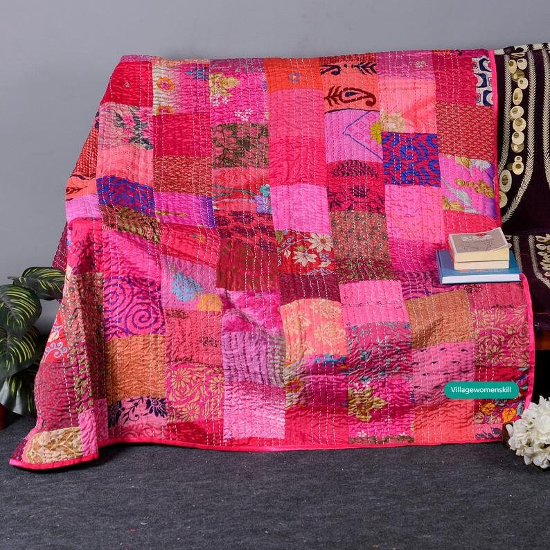 May include: A pink and red patchwork quilt with a variety of floral and geometric patterns. The quilt is made of recycled saris and is hand-stitched. It is a beautiful and unique piece of art that would make a great addition to any home.