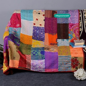 May include: A colorful patchwork quilt made from repurposed saris. The quilt is made up of squares of different colors and patterns, including pink, purple, orange, green, and blue. The quilt is folded and draped over a couch. The text "Villagewomenskill" is visible in the upper right corner of the image.