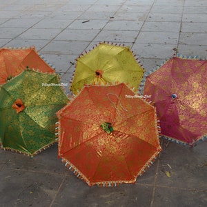 K&ouml;nnte beinhalten: F&uuml;nf bunte dekorative Regenschirme mit goldenen Blumenmustern. Die Regenschirme sind in den Farben Orange, Gr&uuml;n, Gelb und Rosa gehalten. Jeder Regenschirm hat eine dekorative Bord&uuml;re mit Quasten. Die Regenschirme sind ge&ouml;ffnet und auf einer grauen Steinoberfl&auml;che angeordnet.