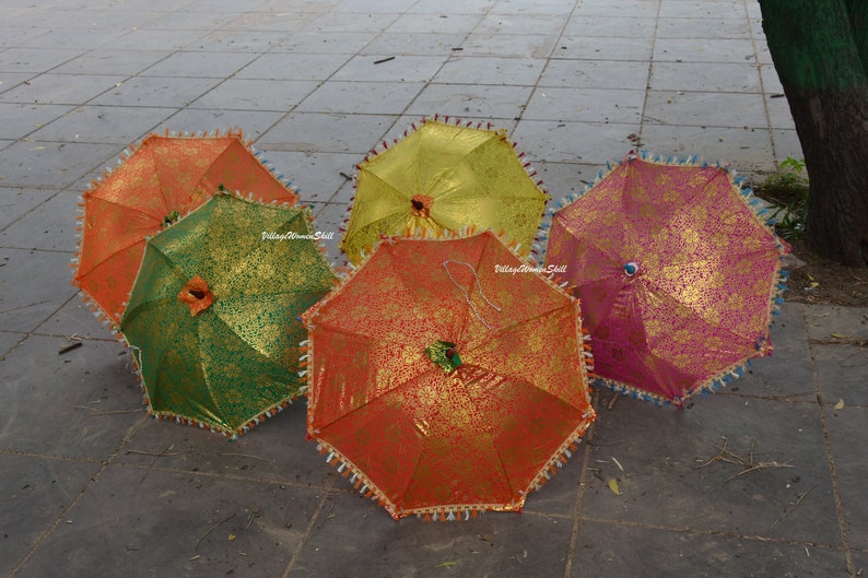 K&ouml;nnte beinhalten: F&uuml;nf bunte dekorative Regenschirme mit goldenen Blumenmustern. Die Regenschirme sind in den Farben Orange, Gr&uuml;n, Gelb und Rosa gehalten. Jeder Regenschirm hat eine dekorative Bord&uuml;re mit Quasten. Die Regenschirme sind ge&ouml;ffnet und auf einer grauen Steinoberfl&auml;che angeordnet.