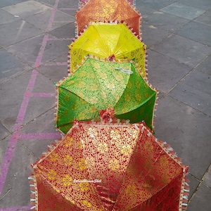 K&ouml;nnte beinhalten: Eine Reihe bunter, dekorativer Regenschirme mit floralen Mustern. Die Schirme sind in Rot-, Gelb- und Gr&uuml;nt&ouml;nen gehalten, mit goldenen Akzenten und Fransen. Sie sind auf einer grauen Oberfl&auml;che angeordnet.
