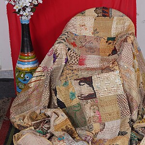 May include: A patchwork quilt made of various fabrics in shades of brown, beige, and cream. The quilt is draped over a chair and has a hand-stitched design. The quilt is made by Village Women Skill.
