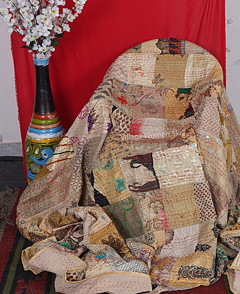 May include: A patchwork quilt made of various fabrics in shades of brown, beige, and cream. The quilt is draped over a chair and has a hand-stitched design. The quilt is made by Village Women Skill.