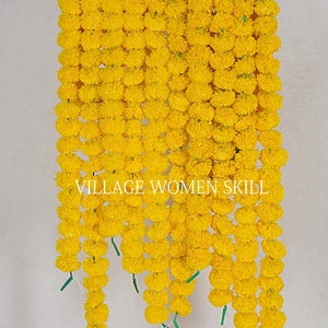 May include: A garland of yellow marigold flowers, strung together in a long row. The garland is hanging from a metal rod, and the flowers are arranged in a symmetrical pattern. The text "VILLAGE WOMEN SKILL" is visible in the center of the image.