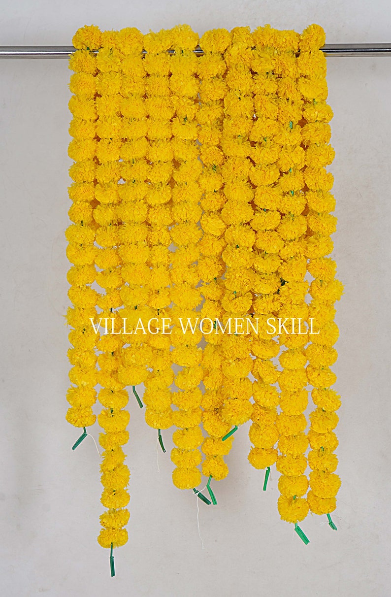 May include: A garland of yellow marigold flowers, strung together in a long row. The garland is hanging from a metal rod, and the flowers are arranged in a symmetrical pattern. The text "VILLAGE WOMEN SKILL" is visible in the center of the image.