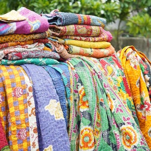 May include: A collection of folded and draped kantha quilts featuring various vibrant colors and intricate patterns. The textiles display floral, geometric, and paisley motifs in shades of yellow, pink, green, blue, and orange with visible stitching.