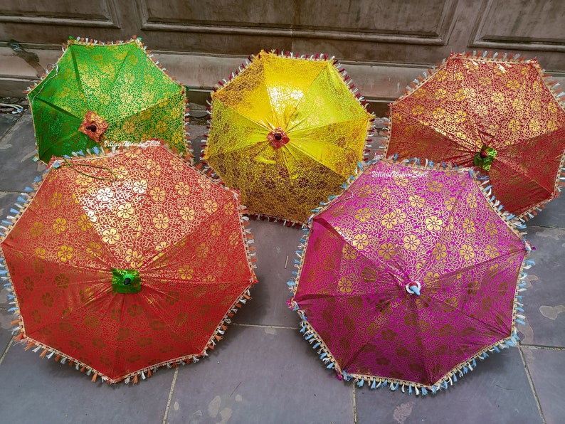 K&ouml;nnte beinhalten: Eine Auswahl an dekorativen Regenschirmen in verschiedenen Farben. Jeder Regenschirm hat ein goldenes Blumenmuster auf einem gr&uuml;nen, gelben, roten und rosa Hintergrund. Die R&auml;nder sind mit Quasten verziert.