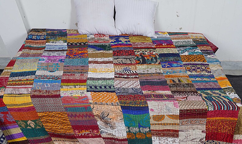 May include: A colorful patchwork quilt made from recycled saris. The quilt is made up of squares of different fabrics and colors, including red, blue, green, yellow, and orange. The quilt is laid out on a bed with white pillows.