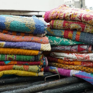 Könnte beinhalten: Ein Stapel bunter Kantha-Steppdecken, hergestellt aus recycelten Saris, mit einer Vielzahl von floralen und geometrischen Mustern. Die Steppdecken sind in einem Stapel angeordnet, wobei die oberste Steppdecke in der Hälfte gefaltet ist. Die Steppdecken sind aus Baumwolle und handgenäht. Copyright@VillageWomenSkill