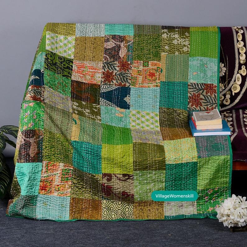 May include: A patchwork quilt made from repurposed saris in shades of green, brown, and blue. The quilt is folded and displayed on a couch with a stack of books and a vase of white flowers. The text "VillageWomenskill" is visible in the lower right corner of the image.