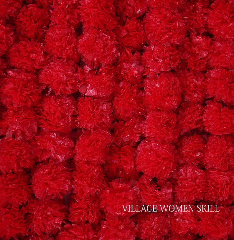 May include: A close-up image of a large quantity of red paper flowers. The flowers are arranged in a dense cluster, creating a vibrant and textured background. The text "VILLAGE WOMEN SKILL" is visible at the bottom of the image.