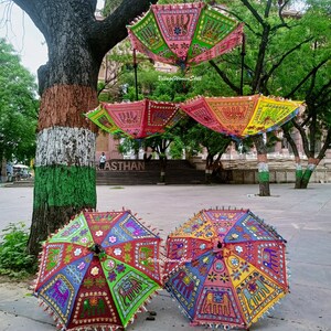 May include: Three colourful umbrellas with intricate embroidery and mirror work, featuring elephants and floral patterns. The umbrellas are hanging from a tree branch and are displayed on a paved surface. The text "Village Women Skill" is visible on one of the umbrellas.