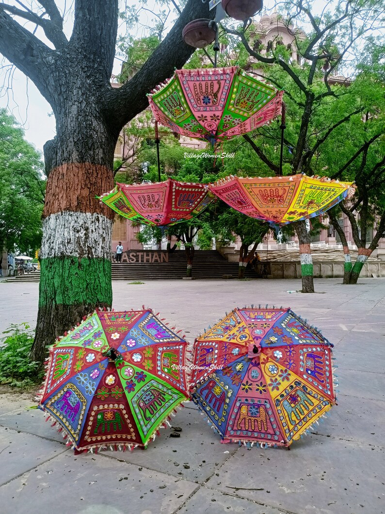 May include: Three colourful umbrellas with intricate embroidery and mirror work, featuring elephants and floral patterns. The umbrellas are hanging from a tree branch and are displayed on a paved surface. The text "Village Women Skill" is visible on one of the umbrellas.