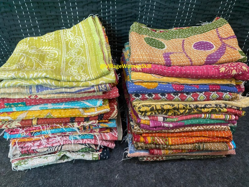 May include: A stack of colorful kantha quilts, made from recycled saris, with intricate patterns and stitching. The quilts are folded and stacked on a dark surface. @VillageWomenSkill