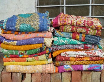 May include: Stacks of colorful, patterned quilts. The quilts feature a variety of designs and colors, including blue, yellow, orange, and pink. The quilts are stacked on a brick surface, with a window in the background.