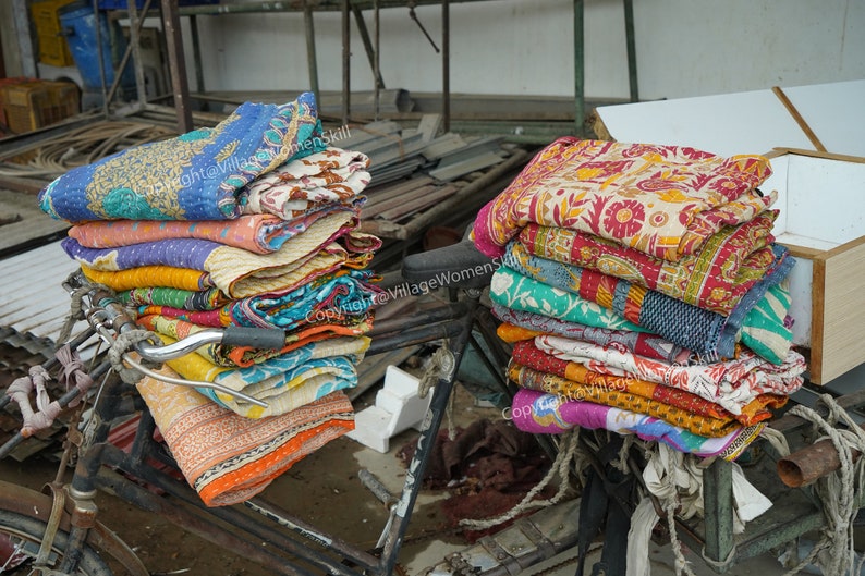 Puede incluir: Una pila de colchas kantha coloridas se apilan en una bicicleta, listas para la venta. Las colchas est&aacute;n hechas de saris reciclados y presentan intrincados patrones y dise&ntilde;os.