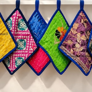 May include: A row of eight square quilted potholders with blue fabric trim and hanging loops. The potholders feature various colours and patterns, including red, yellow, pink, green, purple, and blue, with visible stitching and the text villagewomenskill.