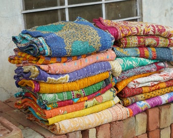 May include: A stack of colorful, patterned quilts. The quilts feature various designs and colors, including blue, yellow, orange, pink, and green. The quilts are folded and stacked on a brick surface.