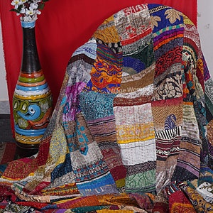 May include: A colorful patchwork quilt made from recycled saris. The quilt is made up of squares of different fabrics, each with a unique pattern and color. The quilt is draped over a chair and has a rustic, vintage look.