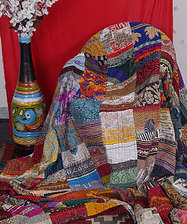 May include: A colorful patchwork quilt made from recycled saris. The quilt is made up of squares of different fabrics, each with a unique pattern and color. The quilt is draped over a chair and has a rustic, vintage look.