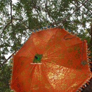 K&ouml;nnte beinhalten: Ein offener, orangefarbener Regenschirm mit goldenem Blumenmuster, verziert mit Quasten, h&auml;ngt an einem Ast. Der Stoff des Schirms hat ein gl&auml;nzendes Aussehen. Der Regenschirm ist mit einer Pfauenfeder verziert.