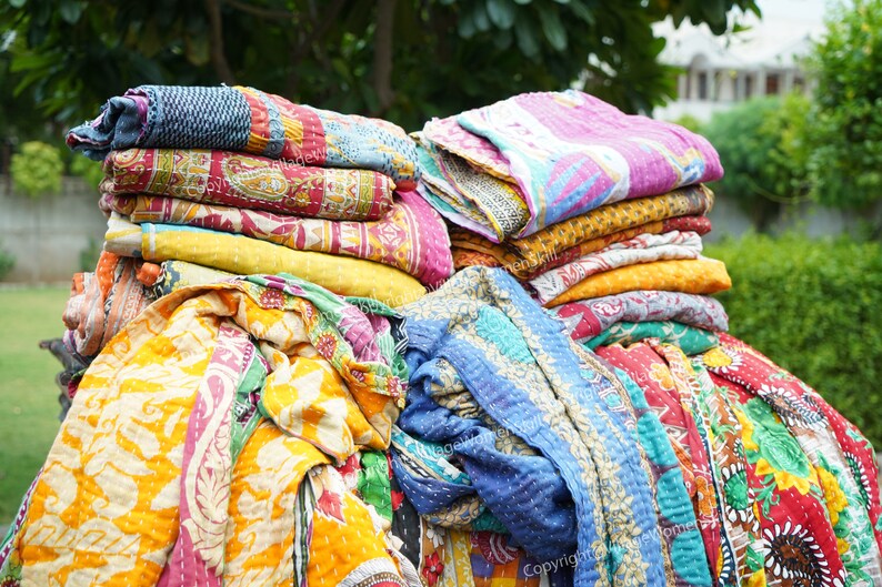 Puede incluir: Una pila de colchas kantha coloridas, hechas de saris reciclados, con una variedad de patrones florales y geom&eacute;tricos. Las colchas est&aacute;n dobladas y apiladas una encima de la otra, creando una exhibici&oacute;n vibrante y texturizada.