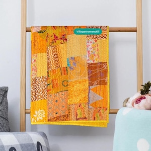 Pu&ograve; includere: Una trapunta patchwork gialla, arancione e marrone con un bordo giallo. La trapunta &egrave; appesa a una scala di legno. Il testo "Villagewomenskill" &egrave; visibile sulla trapunta.