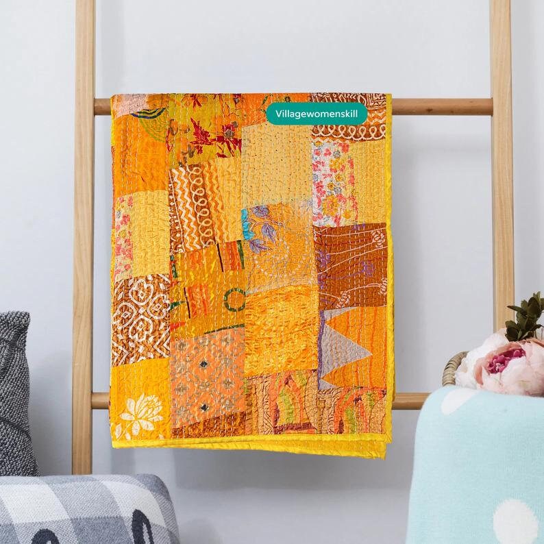 Pu&ograve; includere: Una trapunta patchwork gialla, arancione e marrone con un bordo giallo. La trapunta &egrave; appesa a una scala di legno. Il testo "Villagewomenskill" &egrave; visibile sulla trapunta.