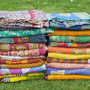 Könnte beinhalten: Ein Stapel bunter Kantha-Steppdecken aus recycelten Saris. Die Decken sind in verschiedenen Mustern und Farben übereinander gelegt, darunter Blau, Grün, Gelb, Orange, Rosa und Lila. Die Decken sind in zwei Stapeln nebeneinander auf einem Bett aus grünem Gras angeordnet.