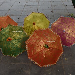 K&ouml;nnte beinhalten: F&uuml;nf farbenfrohe dekorative Regenschirme mit goldenen Blumenmustern. Die Regenschirme sind in den Farben Orange, Gr&uuml;n, Gelb und Rosa gehalten. Jeder Regenschirm hat eine Quastenborte und eine dekorative Spitze. Die Regenschirme sind ge&ouml;ffnet und auf einer grauen Oberfl&auml;che angeordnet.