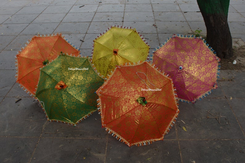 K&ouml;nnte beinhalten: F&uuml;nf farbenfrohe dekorative Regenschirme mit goldenen Blumenmustern. Die Regenschirme sind in den Farben Orange, Gr&uuml;n, Gelb und Rosa gehalten. Jeder Regenschirm hat eine Quastenborte und eine dekorative Spitze. Die Regenschirme sind ge&ouml;ffnet und auf einer grauen Oberfl&auml;che angeordnet.