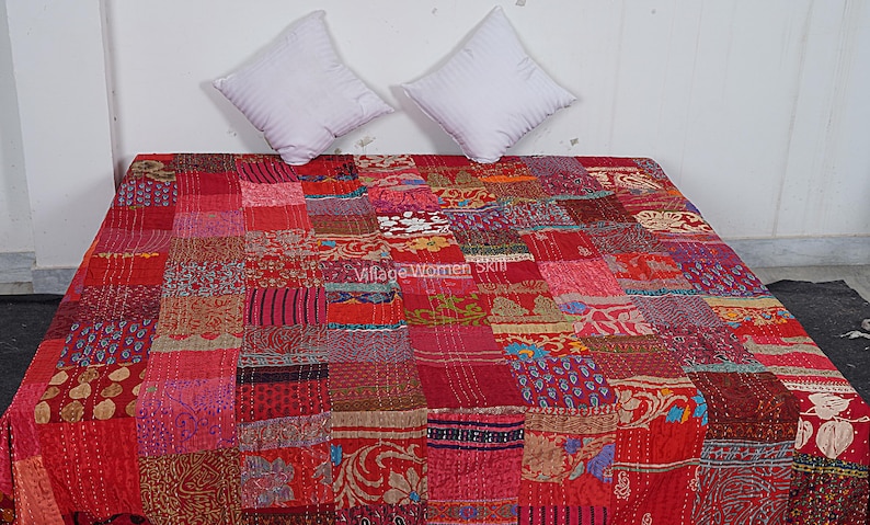 May include: Red and pink patchwork quilt with a variety of floral and geometric patterns. The quilt is made of recycled saris and is hand-stitched. "Village Women Skill" is printed on the quilt.