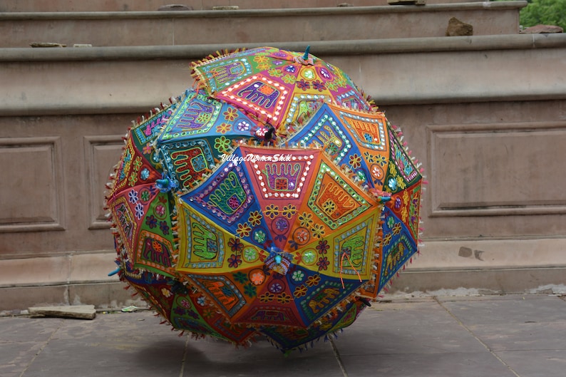 May include: A colourful, round, fabric umbrella with intricate embroidery and mirror work. The umbrella is decorated with geometric patterns, floral motifs, and small mirrors. The umbrella is a vibrant mix of red, orange, yellow, green, blue, and purple.