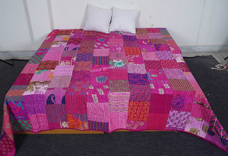 May include: A pink and purple patchwork quilt with a variety of patterns and textures. The quilt is made of recycled saris and is hand-stitched. The quilt is laid out on a bed with two white pillows.
