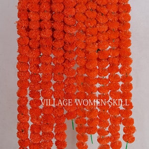 May include: A collection of bright orange marigold garlands suspended against a white backdrop. Each garland is made up of numerous round, fluffy flowers. The text "VILLAGE WOMEN SKILL" is displayed at the bottom of the image.