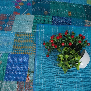 May include: A patchwork quilt with a blue and green color scheme. The quilt is made of different fabrics and has a quilted design. A small potted plant with red berries is in the foreground.