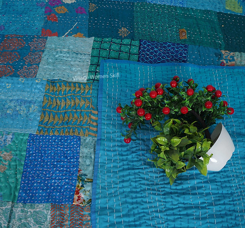 May include: A patchwork quilt with a blue and green color scheme. The quilt is made of different fabrics and has a quilted design. A small potted plant with red berries is in the foreground.